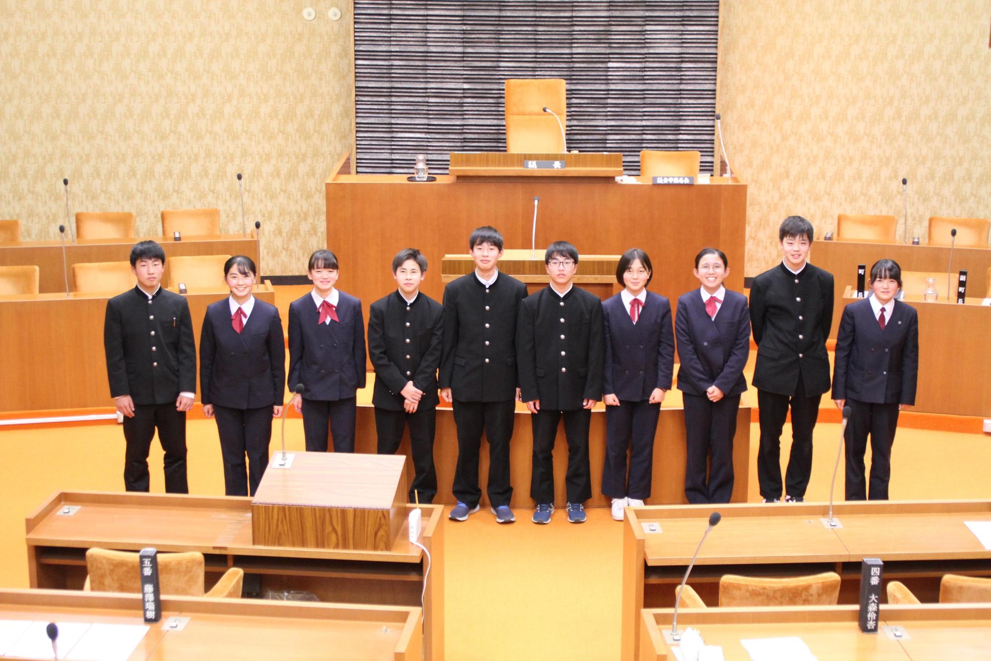 机がたくさん並んでいる辰野町役場の議場で中学生10人が並んで笑顔を向けている写真