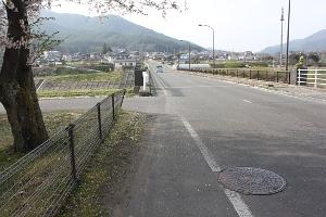左手に桜の木と畑がある広い道路の写真