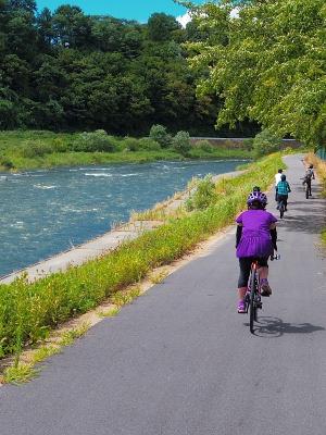 手前の紫のユニフォームに身を包まれ女性の他3人が川沿いの道を自転車に乗って走っている写真