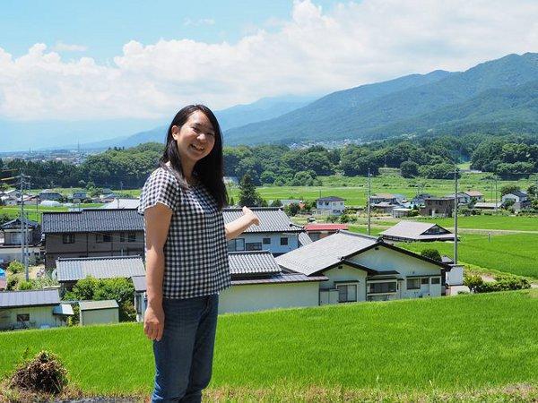 緑色の田山と民家が広がる辰野町を背景に笑顔で立っている女性